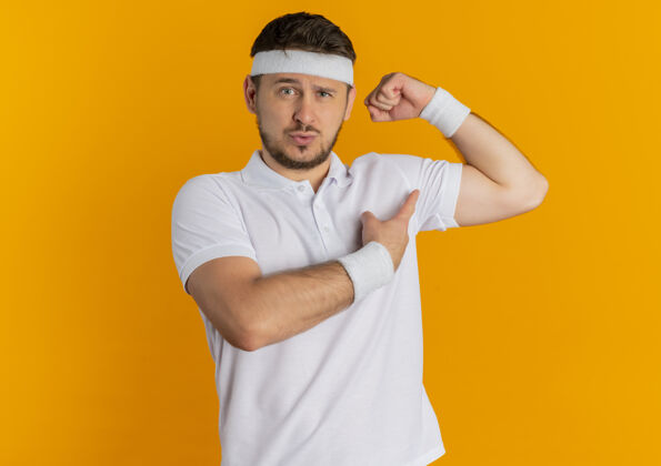 年轻身穿白衬衫 头箍高举拳头的年轻健身男子站在橙色的墙上 两头肌看起来自信而自豪拳头运动装提高