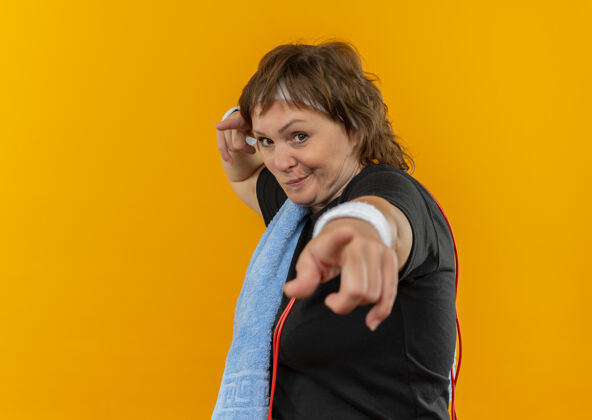 毛巾中年运动女性 身穿黑色t恤 头戴头带 肩上搭着毛巾 手指指着橙色的墙上 带着怀疑的表情运动手指指尖