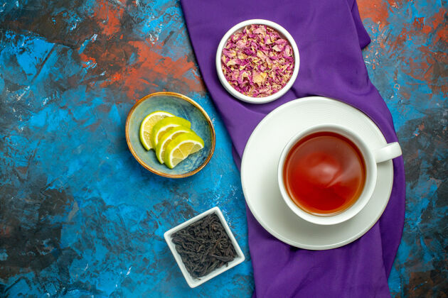 盘子俯视图一杯茶碗 茶碗上放着干花瓣 茶片上放着蓝红色的柠檬紫色桌布桌布早餐干的