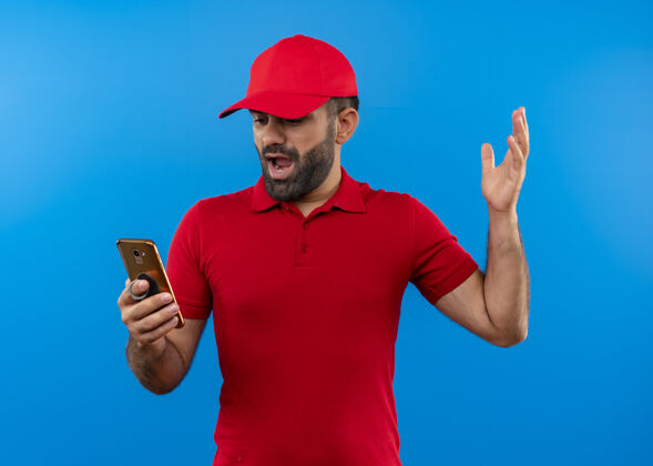 抱着沮丧的留着胡子的送货员穿着红色制服 戴着帽子 举着智能手机 不高兴地站在蓝色的墙上递送举起胡须