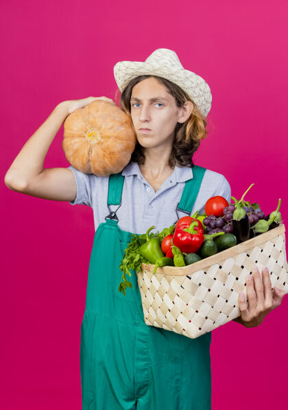 花园年轻的园丁穿着连体衣戴着帽子拿着装满新鲜蔬菜的箱子年轻南瓜持有