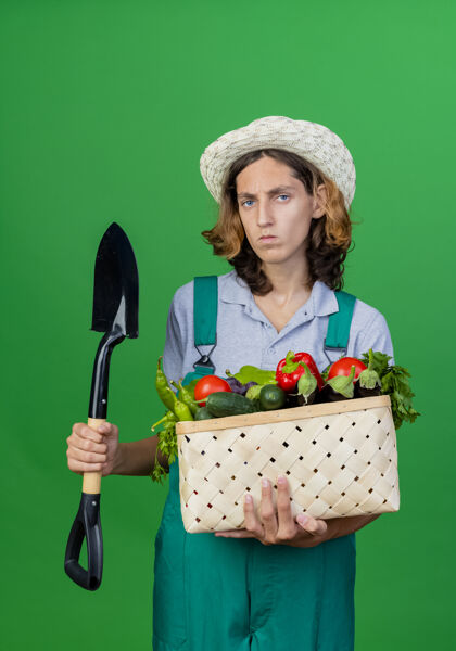 脸年轻的园丁穿着连体衣戴着帽子拿着装满新鲜蔬菜的箱子连身衣年轻蔬菜