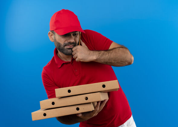 堆着一个留着胡须 身穿红色制服 戴着帽子的送货员站在蓝色的墙上 手里拿着一叠比萨饼盒 一边用手机聊天 一边显得很困惑而说话困惑