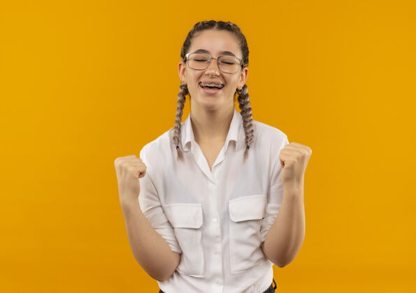 人戴着眼镜 梳着辫子 穿着白衬衫的年轻女学生握紧拳头 高兴而兴奋地站在橙色的墙上为自己的成功而欢呼快乐手势女孩
