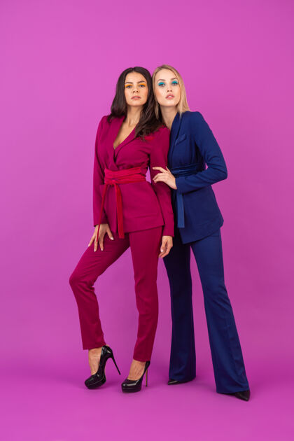 华丽高级时尚风格两位微笑迷人的女士在紫罗兰色的墙壁上穿着时尚多彩的紫色和蓝色晚礼服 朋友们一起玩 时尚潮流秋天奢华衣服