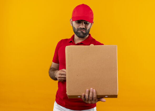 看满脸胡须 身穿红色制服 头戴鸭舌帽的送货员拿着打开的披萨盒 站在橘色的墙上 神情困惑地看着它制服送货困惑