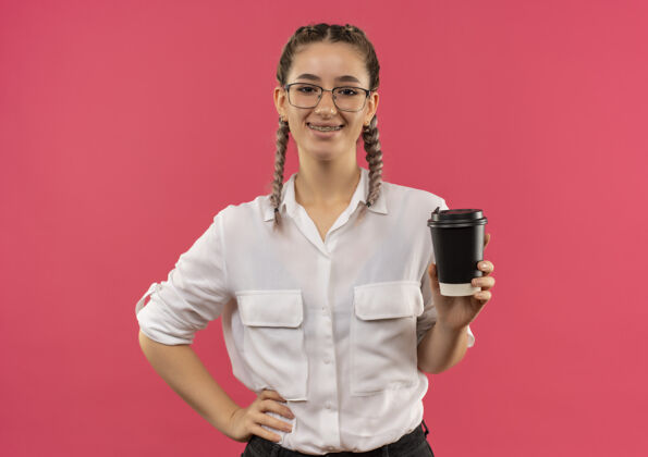 眼镜戴着眼镜 梳着辫子 穿着白衬衫的年轻女学生手里拿着咖啡杯 面带微笑自信地站在粉色的墙上望着前方学生姿势微笑