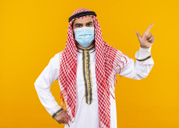 手指阿拉伯商人穿着传统服装 戴着护面面具 自信的表情指着侧面 食指站在橙色的墙上指信心面部