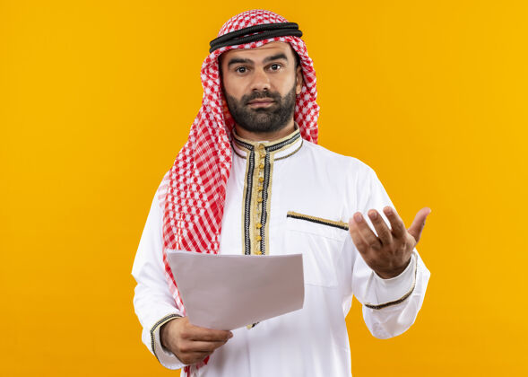 传统身着传统服饰的阿拉伯商人站在橙色的墙上 手拿着文件 伸出胳膊问问题文件站着手臂