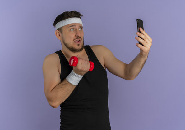 运动员戴着头巾的年轻健身男子用智能手机自拍 手里拿着哑铃 站在紫色的墙上 看上去很困惑采取手移动