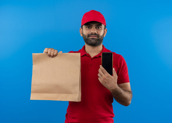 制服留着胡须的送货员穿着红色制服 戴着帽子 手里拿着纸包 微笑着把智能手机放在蓝色的墙上展示站着男人
