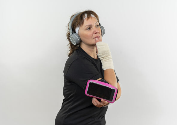 表情中年运动女性 身穿黑色t恤 头戴头带 手戴智能手机臂章 面带沉思的表情 站在白墙上思考女人臂章移动