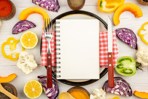 早餐俯视图食谱写在记事本上刀叉放在圆板上切蔬菜不同的香料放在碗里放在白色的木桌上蔬菜切割叉子