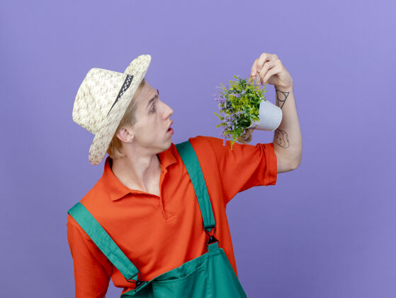 穿年轻的园丁穿着连体衣戴着帽子拿着盆栽植物惊讶地看着它连身衣站帽子