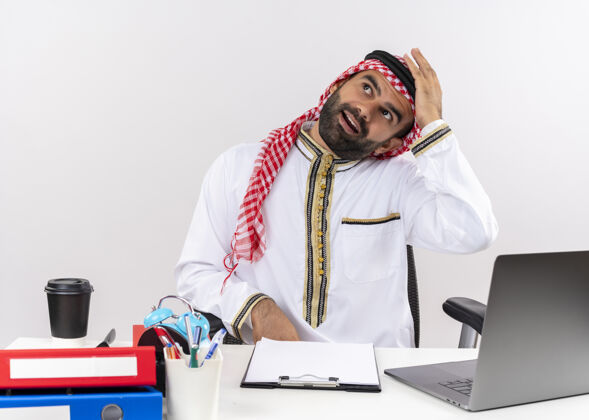 工作身着传统服装的阿拉伯商人坐在桌旁 拿着笔记本电脑 神情迷茫 在办公室工作时有疑虑穿阿拉伯语传统