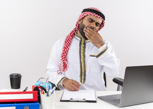 笔记本电脑身着传统服装的阿拉伯商人坐在桌旁 手提电脑看起来很累 想睡觉打呵欠在办公室工作电脑工作睡眠