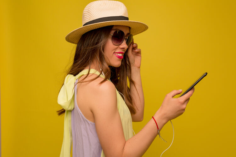 皮肤好幽默的黑发女人在正确的夏装和帽子在黄色墙上使用智能手机活力学生兴奋