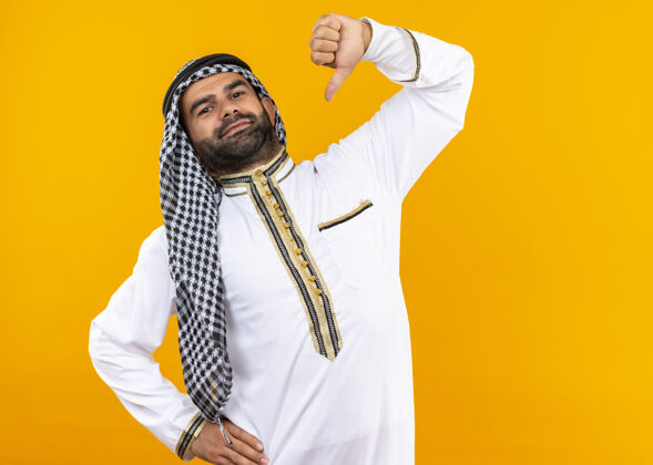 感觉身着传统服饰的阿拉伯商人指着自己自鸣得意的感觉自豪地站在橙色的墙上自豪传统穿