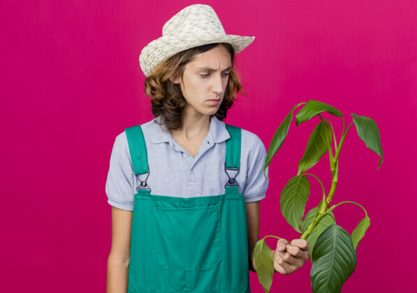 连身衣年轻的园丁穿着连体衣戴着帽子拿着植物严肃地看着它园丁穿着年轻