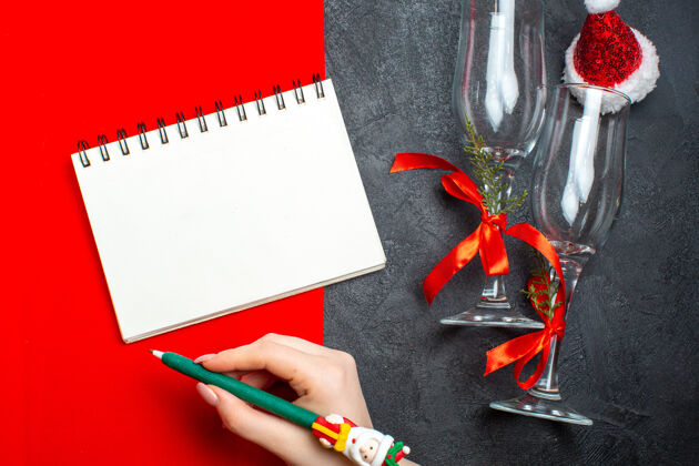 顶部螺旋笔记本俯视图和手持一支笔旁边的玻璃高脚杯圣诞老人帽子的红色和黑色背景书写工具圆珠笔钢笔