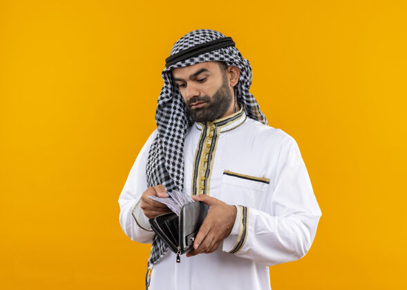 站着身着传统服装的阿拉伯商人站在橙色的墙上 严肃地看着钱包里的现金传统穿看
