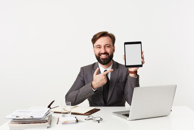 笔记本快乐英俊的黑发年轻人 胡须 穿着正式的衣服 举着平板电脑坐在桌旁 面带微笑 愉快地看着前面工作商务人士深色