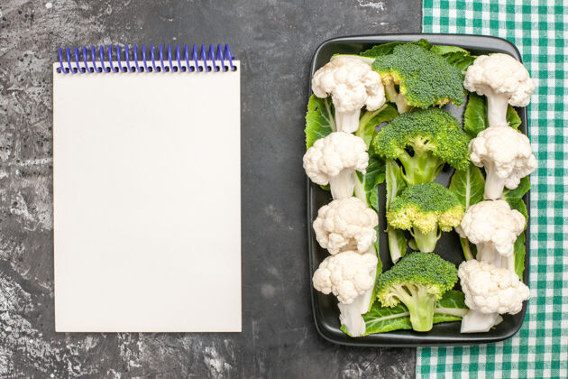 花椰菜在绿色和白色方格餐巾上的黑色矩形板上俯视生西兰花和花椰菜在黑暗的表面上的笔记本西兰花餐巾纸药草
