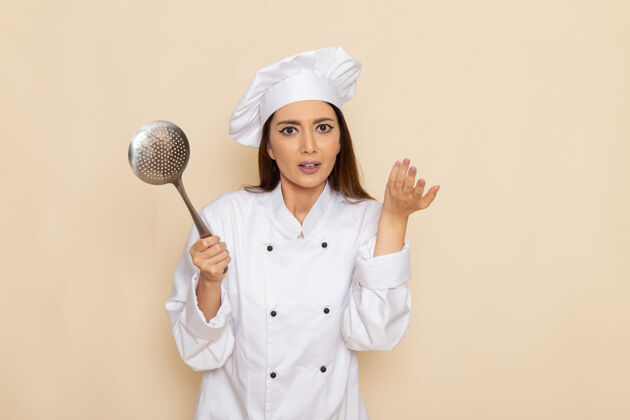 前面身穿白色厨师服的年轻女厨师正拿着大银勺站在白色的墙上比赛球员烹饪