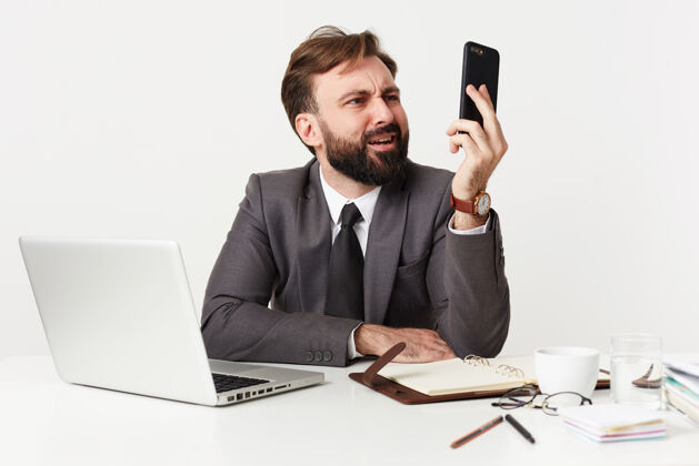 胡须不高兴的年轻黑发男子留着胡子 留着时髦的发型 穿着正式的衣服 一边拿着笔记本电脑和笔记本电脑在办公室工作 一边撅着嘴看着智能手机商务人士发型不高兴