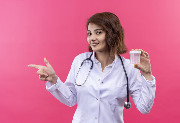 听诊器穿着白大褂的年轻女医生拿着听诊器 微笑着 自信地用手指着一边手指抱着站着