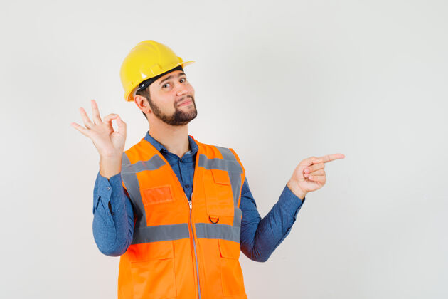 杂工年轻的建设者展示ok标志 指着一边穿着衬衫 背心 头盔 看起来很高兴前视图工人工业制服