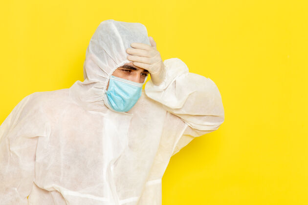 浴袍穿着白色特殊防护服 带无菌口罩的男性科学工作者的正面图 黄色墙壁上有头痛套装头痛长袍