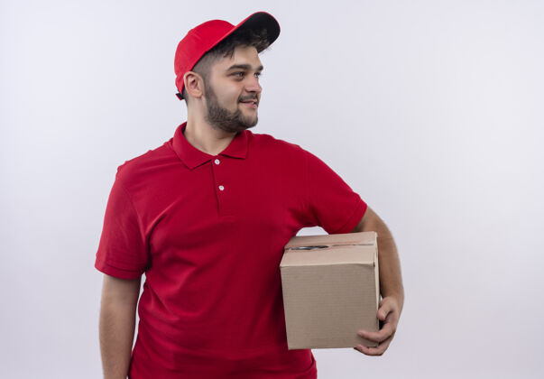 男人年轻的送货员穿着红色制服 拿着帽子的盒子包 微笑着自信地看着一旁微笑盒子包装