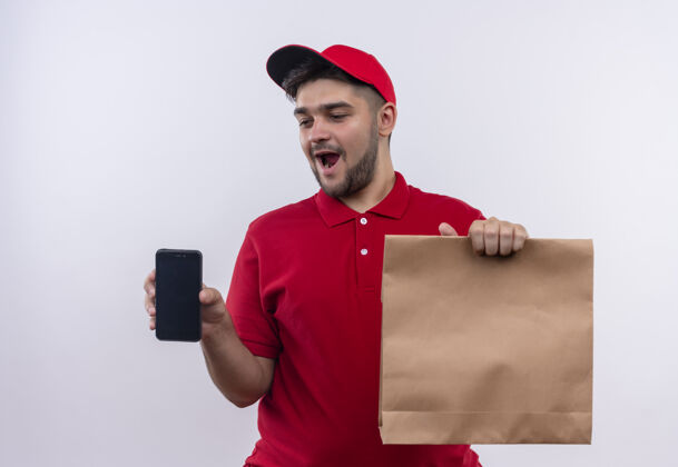 包装年轻的送货员身穿红色制服 戴着帽子 手里拿着纸包 兴高采烈地笑着展示智能手机男孩红色表演
