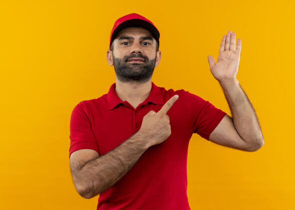 手臂有胡子的送货员穿着红色制服 戴着帽子 手指着胳膊站在橙色的墙上交货手指看