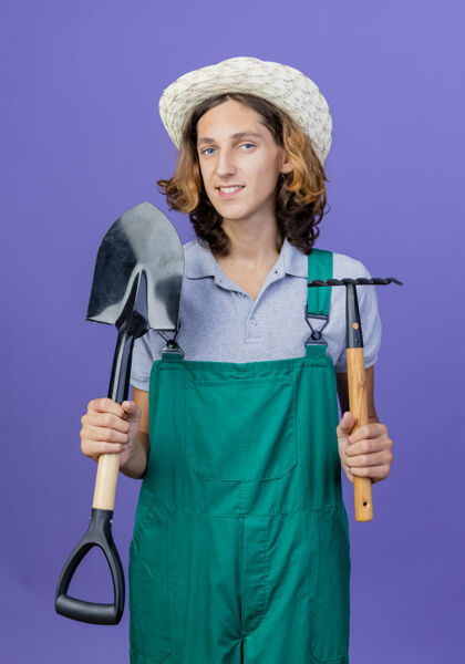 穿年轻的园丁 穿着连体衣 戴着帽子 手里拿着小耙子和铲子站着铲子微笑