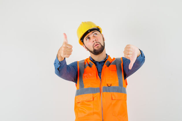 保护身穿衬衫 背心 头盔的年轻建筑工人上下竖起大拇指 神情犹豫不决 俯视前方羽绒服肖像建筑