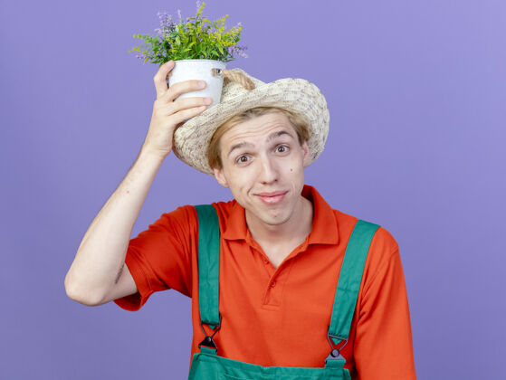 帽子年轻的园丁 穿着连体衣 戴着帽子 头上抱着盆栽植物 微笑着男人连身衣穿着