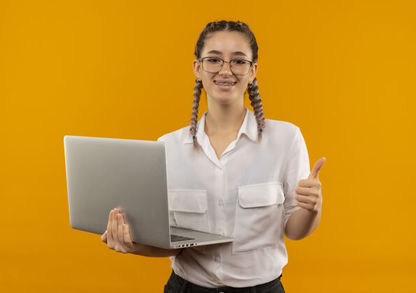 拇指戴着眼镜 梳着辫子 穿着白衬衫的年轻女学生拿着笔记本电脑 面带微笑地站在橙色的墙上竖起大拇指年轻人笔记本电脑人