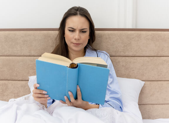 书穿着蓝色睡衣躺在床上的年轻漂亮女人严肃坐着床