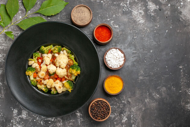 晚餐俯瞰西兰花和花椰菜色拉在黑色椭圆形盘子上不同的香料在小碗的深色表面自由的地方烤箱地方炊具