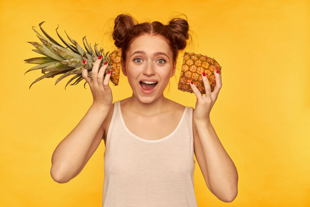 雀斑十几岁的女孩 快乐的红发女人 带着两个发髻穿着白衬衫 把切好的菠萝放在脸旁 健康的生活方式隔着黄色的墙看着孤独健康成人切割