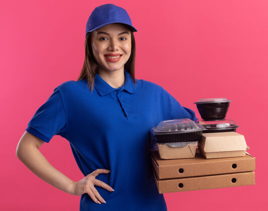 漂亮身着制服 面带微笑的漂亮送货员把手放在腰上 拿着比萨饼盒上的食品包和容器 这些比萨饼盒被隔离在粉红色的墙上 并留有复印空间腰食物女人