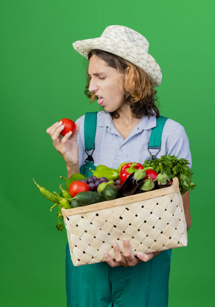 满的年轻的园丁穿着连体衣戴着帽子拿着装满新鲜蔬菜的箱子年轻恶心表情