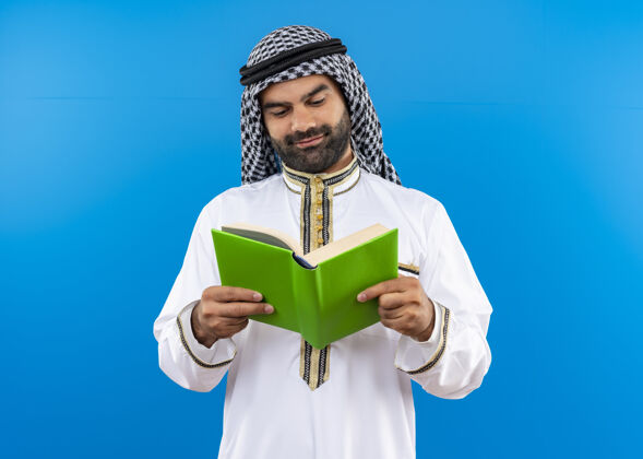 阿拉伯语身着传统服饰的阿拉伯商人手持一本打开的书 面带微笑地站在蓝色的墙上看着它看站商务人士