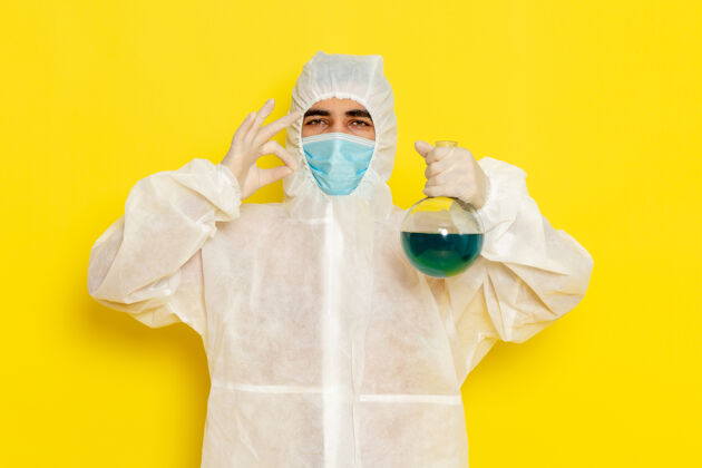 专业身穿特殊防护服的男性科学工作者的正面图 黄色墙壁上有面罩固定瓶医学医生手持