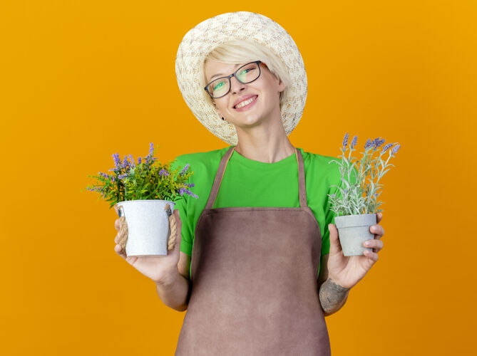欢呼围裙上留着短发 戴着帽子 手里拿着盆栽植物的年轻女园丁站在橙色的背景下看着镜头 开心地微笑着帽子立场头发