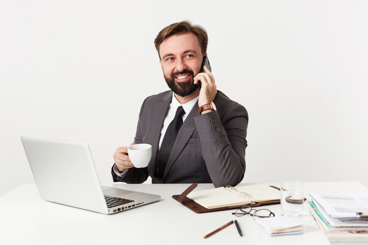 衬衫摄影棚拍摄的是一位留着胡须 留着短发的男性 他正坐在工作桌前 端着一杯咖啡 用智能手机打电话 脸上露出灿烂的笑容爱未剃须理发