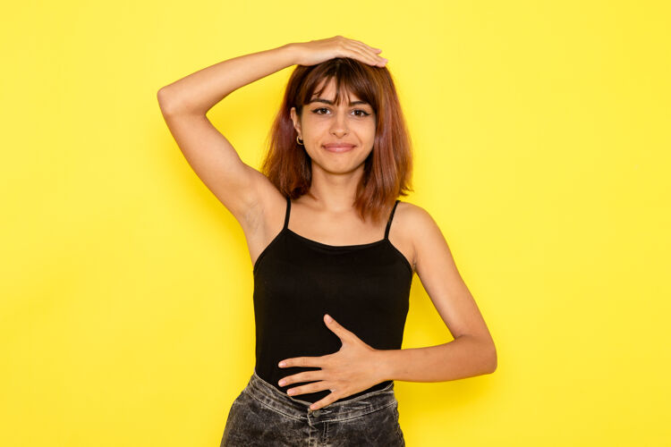 年轻身穿黑色衬衫和灰色牛仔裤的年轻女性在黄色墙壁上微笑和摆姿势的正面视图衬衫情绪微笑