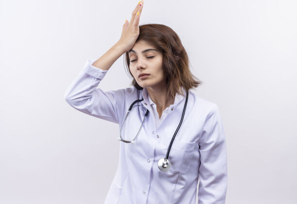 手穿着白大褂 戴着听诊器的年轻女医生看起来很累 手放在头上 眼睛闭着 工作过度外套听诊器医生